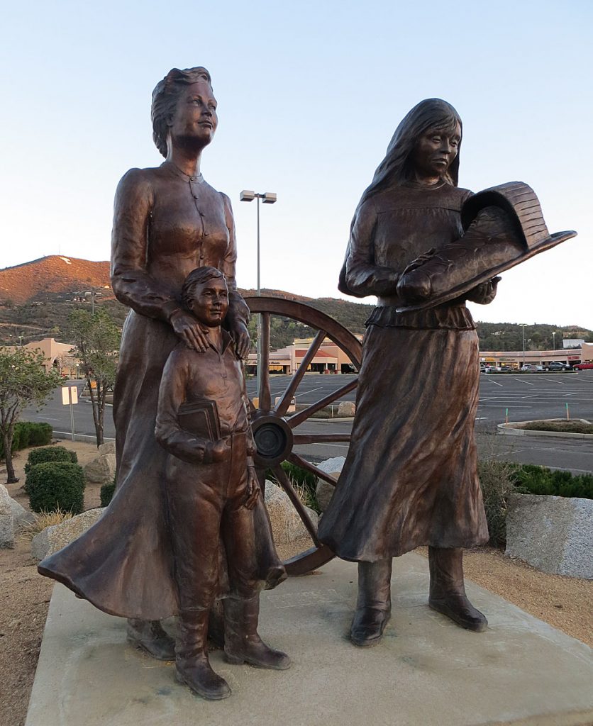 Statue of woman in long dress with young boy and woman holding cradleboard. A wagon wheel stands behind them