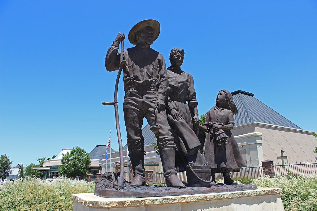 Bronze statue of a man holding a farm tools, a woman, and a young girl holding a rag doll. A bucket sits at their feet.