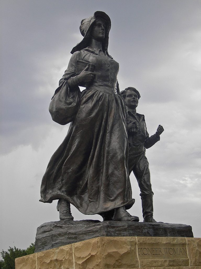Bronze statue of a woman in a sunbonnet striding confidently. She carries a Bible and satchel in her proper right arm. On her left strides a young boy.