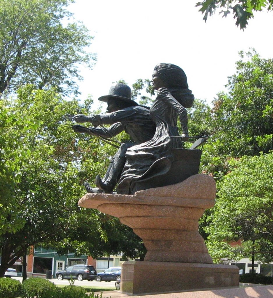 Bronze statue of a man and woman sitting on a wagon box. The man's arms are in front of him as if holding reins.